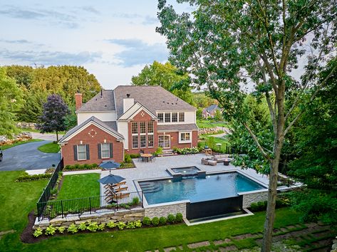 Isn't she lovely? Dominion features a negative edge, spa with an overflow, and dark grey pool tile transforming this traditional rectangular pool into a jaw-dropping work of art. With its dark, moody water and dramatic overflow cascading down a stone veneer retaining wall, the space feels sophisticated and opulent with a pool as beautiful as it is functional. Pool On Sloped Yard Retaining Walls, Inground Pool And Fire Pit Ideas, Pool With Wall On One Side, Pool Perpendicular To House, Red Brick House With Pool, Sloped Yard Pool, Grey Pool Tile, Mobile Fire Pit, Raised Swimming Pool