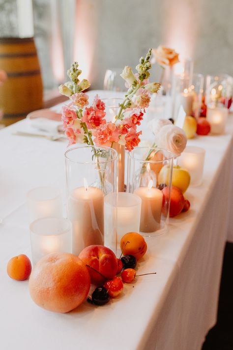 Using the colors of a peach as inspiration, this sweetheart table was filled with neutral colors so the brightness of the fruit and flowers stood out.  Taupe candles, colorful flowers and fruit across the front of the sweetheart table. Fruit Inspired Wedding Decor, Peach Fruit Wedding, Wedding Table With Fruit, Wedding Centerpieces With Fruit, Fruit On Wedding Tables, Fruit Wedding Table Decor, Table Decor With Fruit, Fruit Wedding Centerpieces, Peach Table Decor