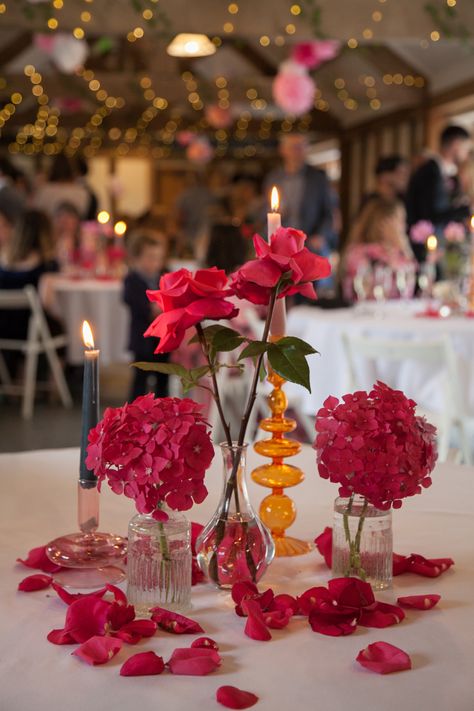 Round table decor in a barn venue. Round table with a white tablecloth. Pink and orange contemporary glass candlesticks with coloured candles. Single stem pink hydrangeas and pink roses in bud vases. Pink rose petals are scattered around. Single Vase Centerpiece Wedding Ideas, Pink And Orange Bud Vases, Red And Pink Wedding Table Decor, Pink And Orange Wedding Centerpieces, Single Stem Table Decor, Pink Bud Vases Wedding, Hydrangea Bud Vase, Hot Pink And Orange Party Decorations, Single Stem Centerpiece