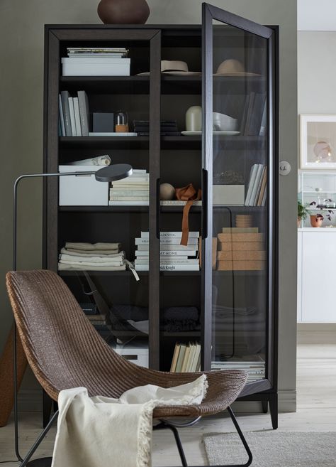 An IKEA REGISSÖR black brown glass door cabinet filled with boxes, books and glassware, behind a chair. Ikea Regissor, Ikea Glass Door Cabinet, Shelf Cover, Modern Home Offices, Black Bookcase, Bookcase With Glass Doors, Bookshelves In Living Room, Glass Front Cabinets, Ikea Cabinets