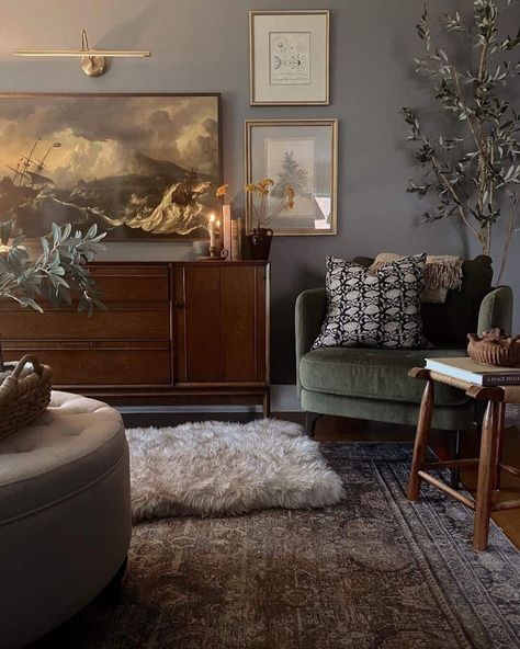 A distinguished essence is displayed within this living room, where earth tones and vintage furniture create a warm atmosphere. Gray walls welcome the company of framed images and a linear sconce. A vintage rug lays over dark wood flooring and hosts a circular ottoman and upholstered armchair...   Image: steph.homebody Room Ideas With Dark Wood Furniture, Dark Earth Tone Living Room, Dark Flooring Living Room, Living Room Dark Wood Floor, Vintage Living Room Aesthetic, Moody Vintage Living Room, Dark Wooden Floor Living Room, Moody Neutral Living Room, Earth Living Room