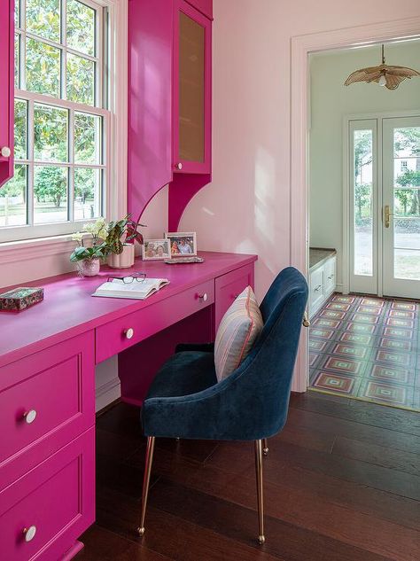 A dark blue velvet chair sits at a hot pink built-in desk fixed beneath a window flanked by glass front hot pink upper cabinets. Hot Pink Bathrooms, Blue Velvet Chair, Den Library, Pink Velvet Chair, Blue Velvet Chairs, Pink Rooms, Dark Blue Velvet, Library Office, Pink Desk