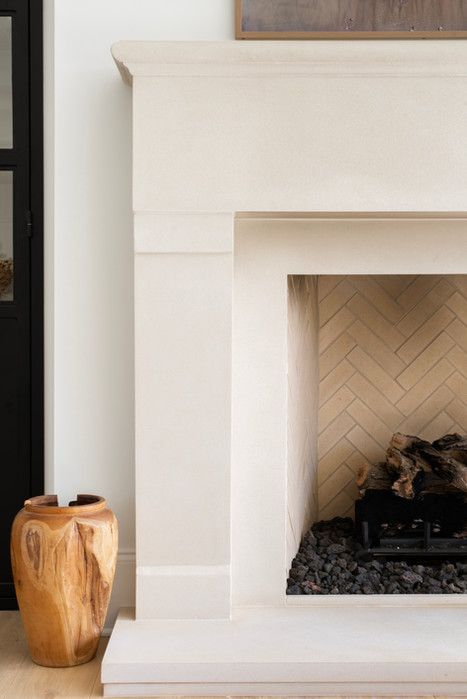 A limestone fireplace mantel and surround detail showcases the fireplace leg profiling and fireplace mantel shelf detail. Clean, simple lines pervade this contemporary fireplace mantel design, which features a cream limestone finish. The fireplace hearth is slightly elevated above the white oak floors, and a decorative wood-carved vase sits next to the fireplace leg on its left. The firebox features a herringbone pattern firebrick and black pebbles that line the bottom of the fireplace. Cream Limestone, Cast Stone Fireplace Surround, Limestone Fireplace Surround, Limestone Mantel, Fireplace Design Ideas, Cast Stone Fireplace, Stone Fireplace Mantel, Stone Fireplace Surround, Limestone Fireplace