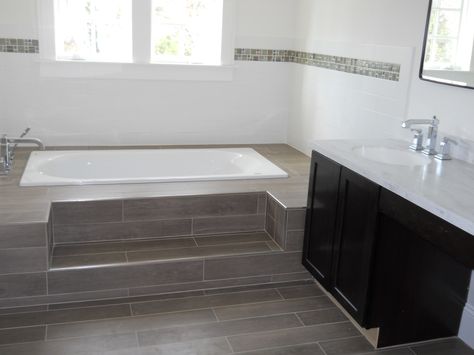 A full bathroom in the Fair Haven house has a bathtub surrounded by long dark grey tiles that match the floor. The step-up bathtub is adjacent to the double sink counter. Built In Bathtub Tub Surround, Bathtub Tile Surround, Sink Counter, Bathtub Surround, Built In Bathtub, Fair Haven, Built In Bath, Drop In Tub, Bathtub Tile