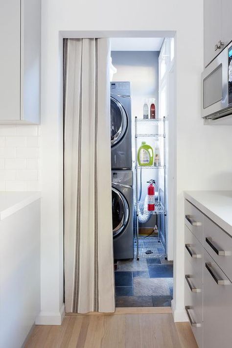 Separated from the kitchen by a gray curtain hung from a doorway, a modern laundry room boasts a stacked silver front loading washer and dryer positioned beside a stainless steel industrial shelving unit. Doors To Hide Washer And Dryer, How To Hide Laundry Machines, How To Hide Washer And Dryer In Hallway, Washer And Dryer In Closet Small Spaces, Hide Washer And Dryer In Kitchen, How To Hide Washer And Dryer In Kitchen, Hiding Washer And Dryer, Curtains To Hide Washer And Dryer, Hide Washer And Dryer