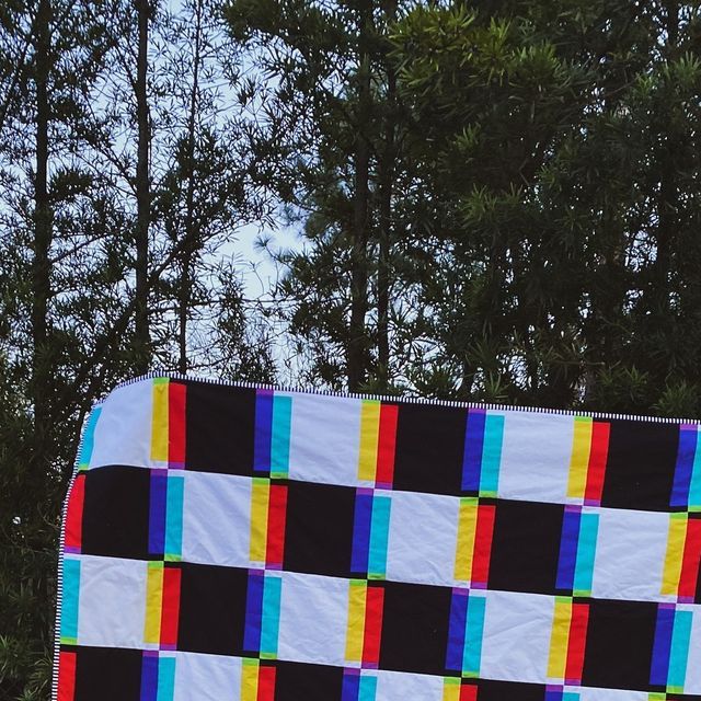 a large multicolored checkered quilt is hanging from a clothes line with trees in the background