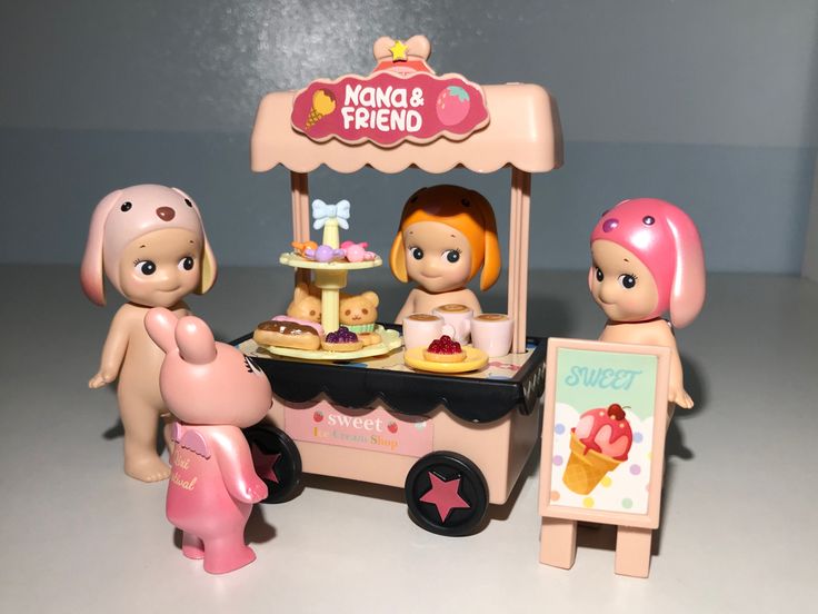 a small toy ice cream cart with three little dolls standing next to it and one is holding a sign that says kona & friend