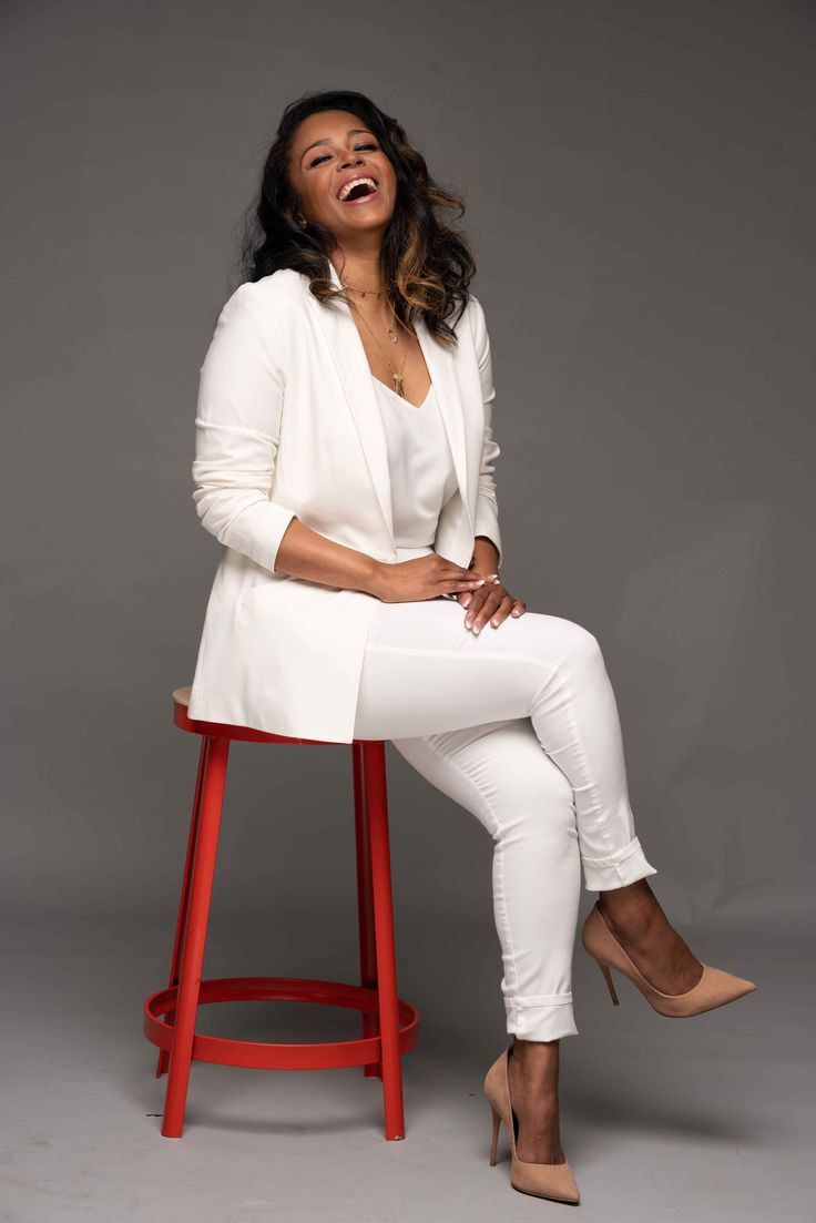 a woman sitting on top of a red stool in white pants and blazer smiling