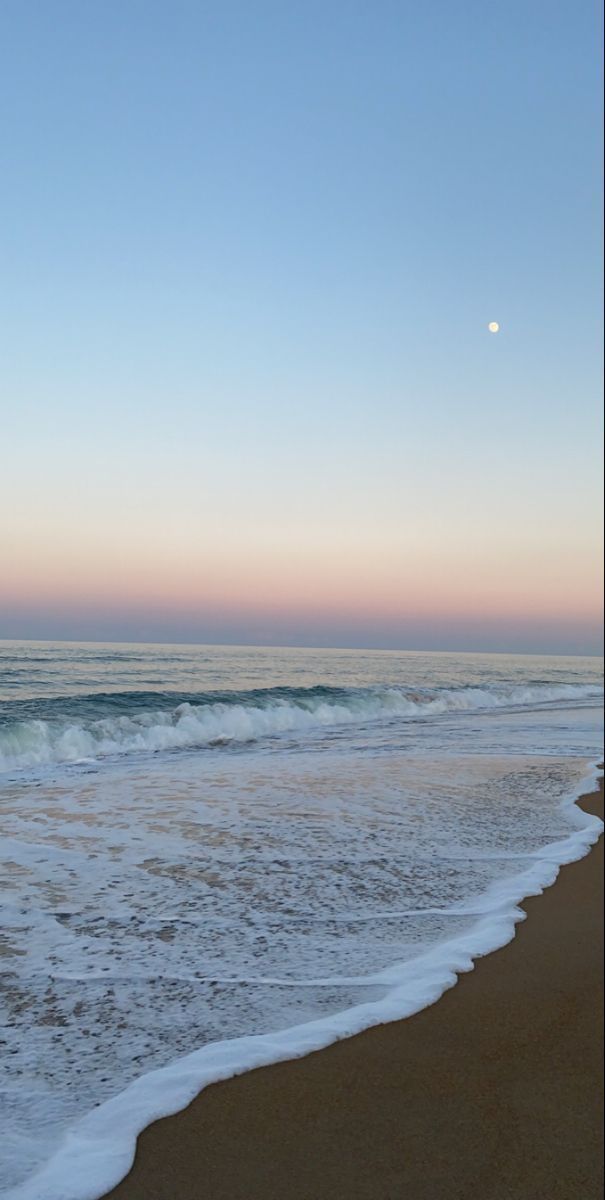 the ocean waves are rolling in to shore at sunset with a half moon rising over the horizon