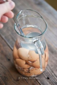 a glass pitcher filled with liquid sitting on top of a wooden table next to a person's hand