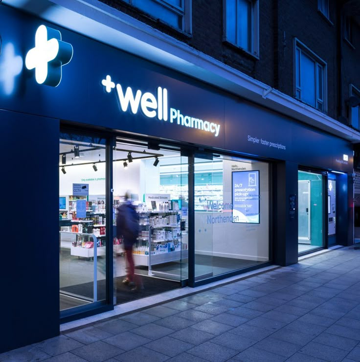 a store front with the words well pharmacy lit up at night, and people walking by