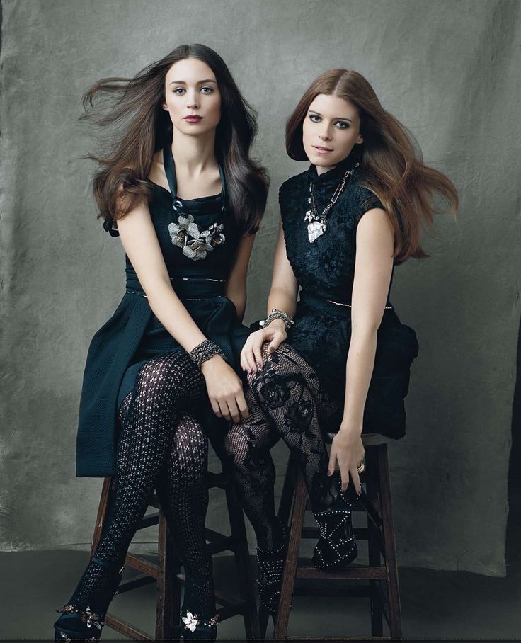 two beautiful young women sitting next to each other on top of a wooden chair in front of a gray background