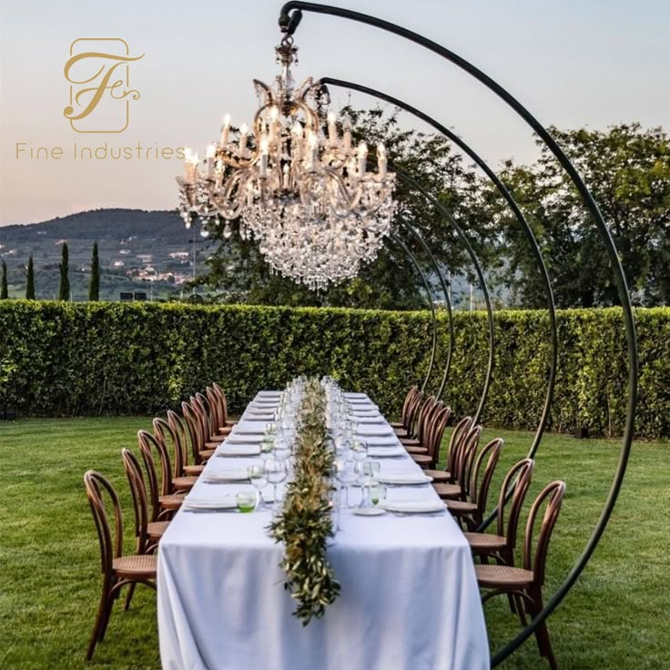 a long table is set up in the middle of a lawn with chandelier hanging from it