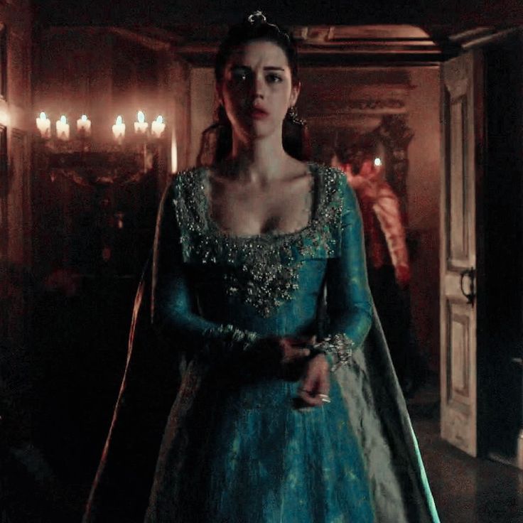 a woman in a blue gown and tiara standing in a dark room with candles on the walls