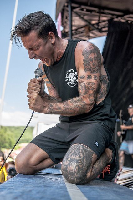 a man sitting on top of a stage with a microphone in his hand and tattoos on his arm