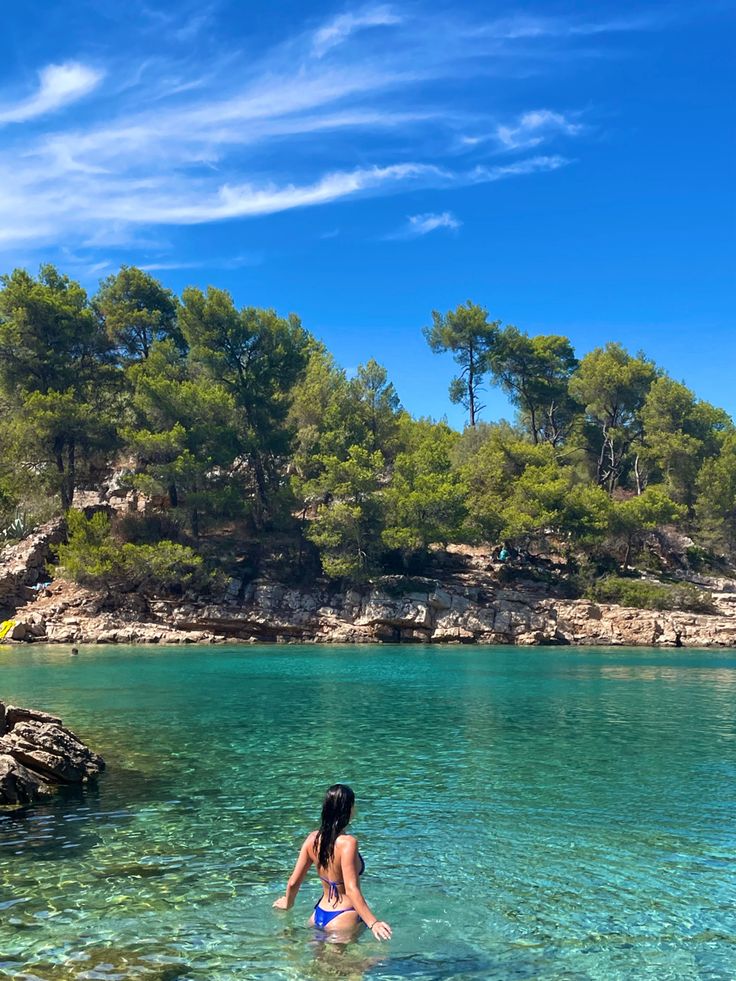 a woman wading in the clear blue water on a sunny day with trees behind her