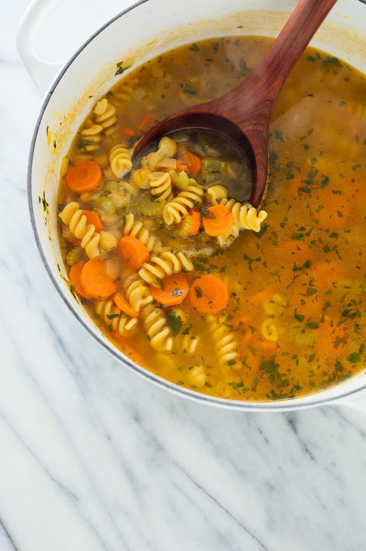 a wooden spoon in a pot filled with noodles and carrots, next to a marble counter top