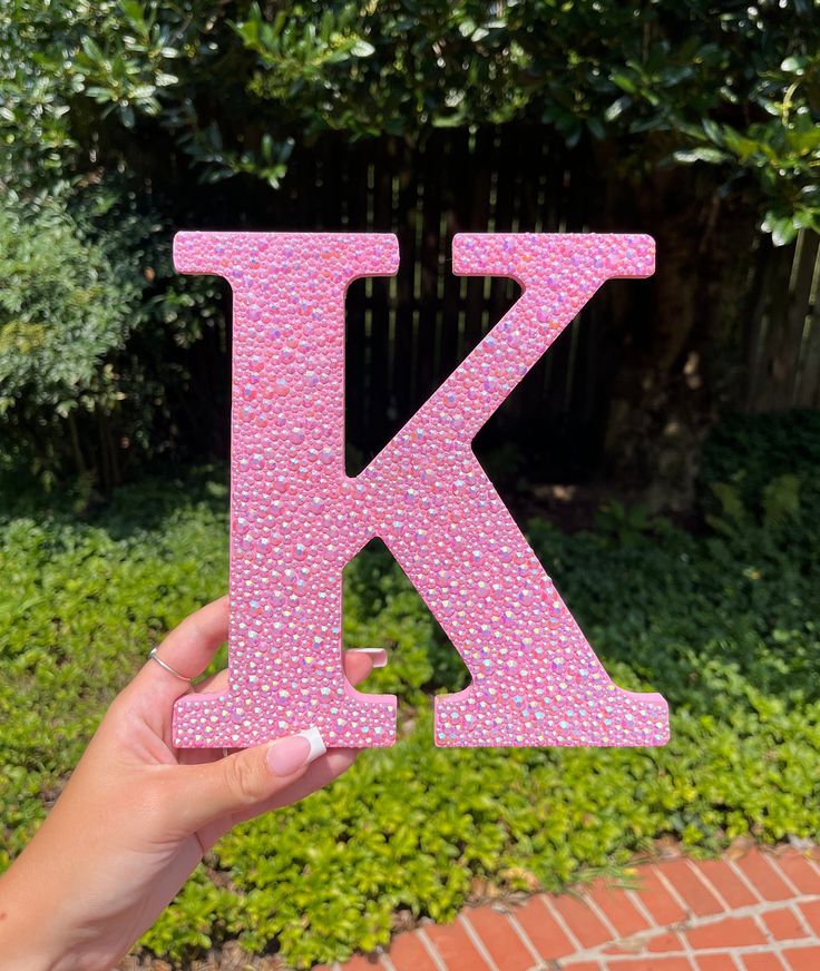 a person holding up a pink letter that is shaped like the letter k in front of some bushes