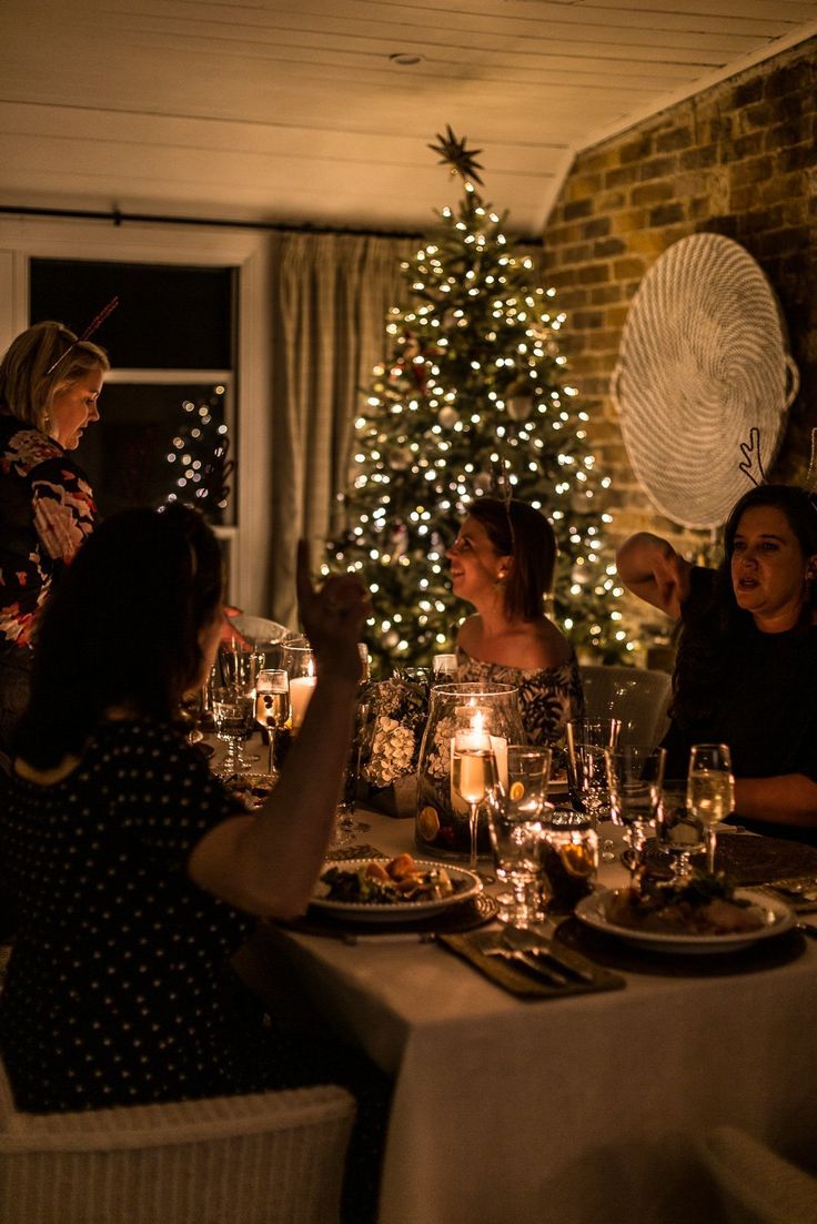 a group of people sitting around a table with food and drinks in front of a christmas tree