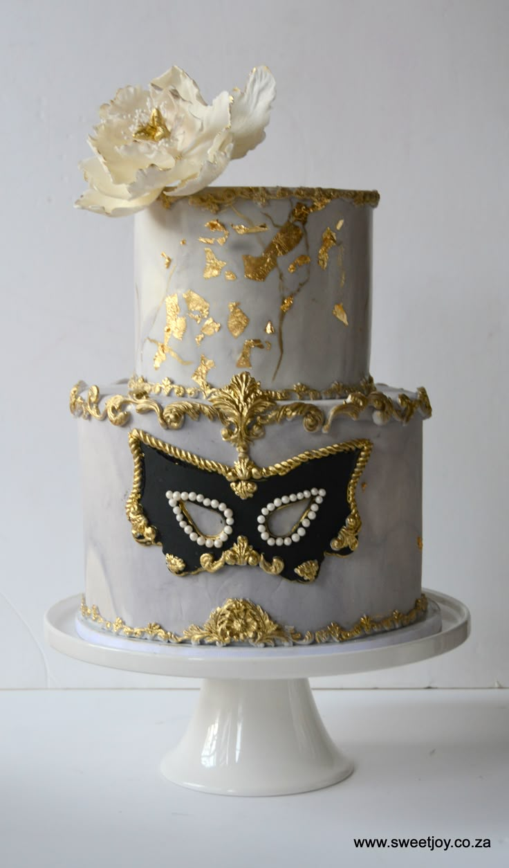 a three tiered cake decorated with gold and black masquerades on a white pedestal