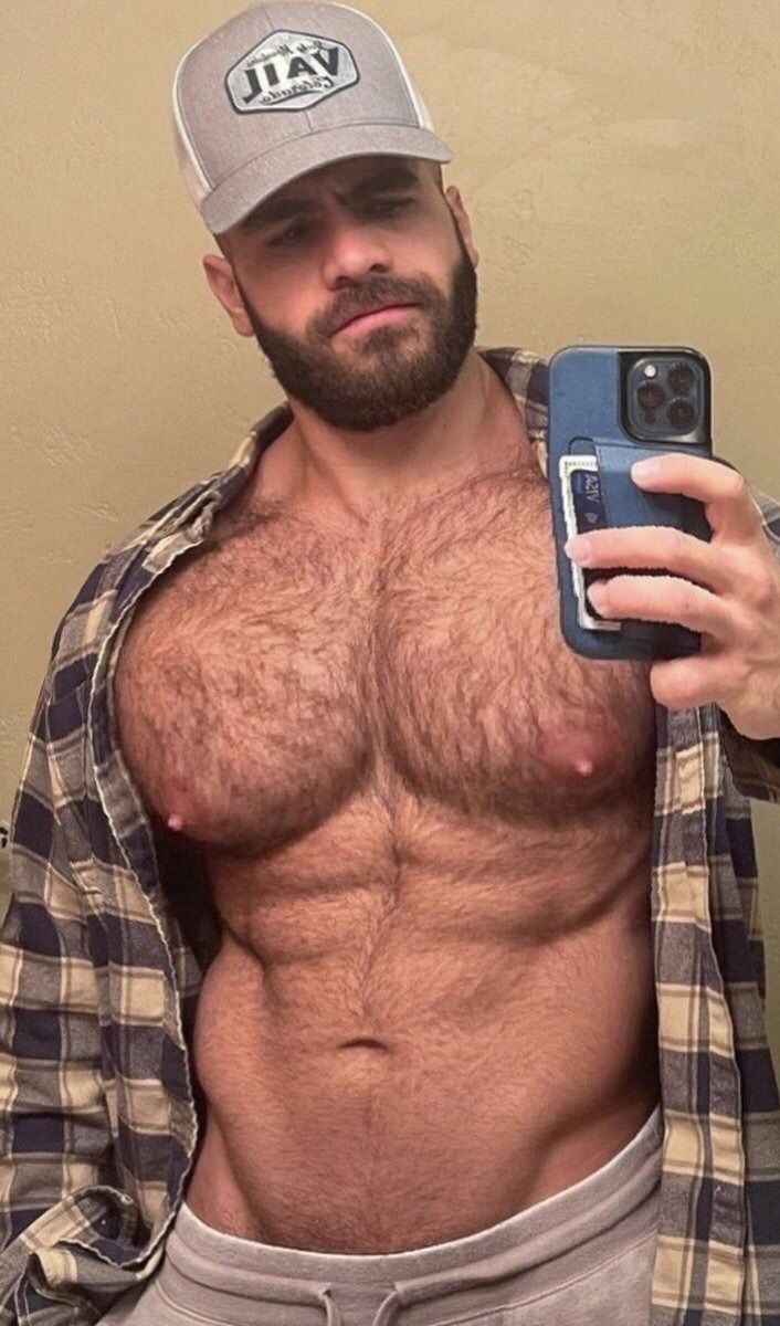 a shirtless man taking a selfie with his cell phone and wearing a hat