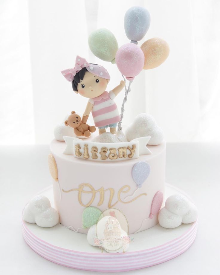 a birthday cake decorated with balloons and a girl holding a teddy bear on the top