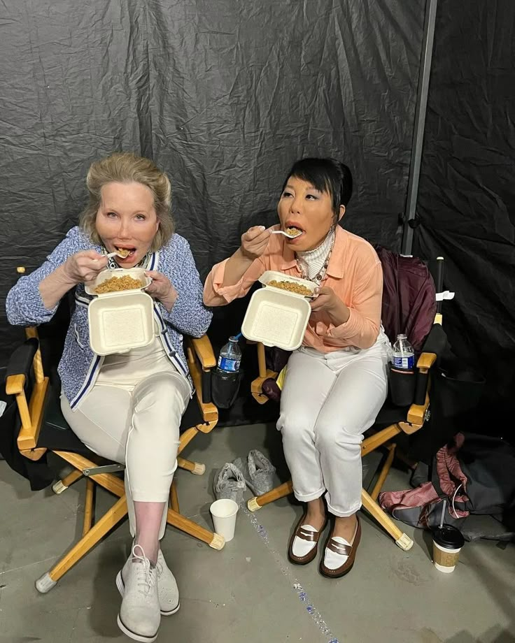 two women sitting in chairs eating food with their mouths open and one holding a sandwich