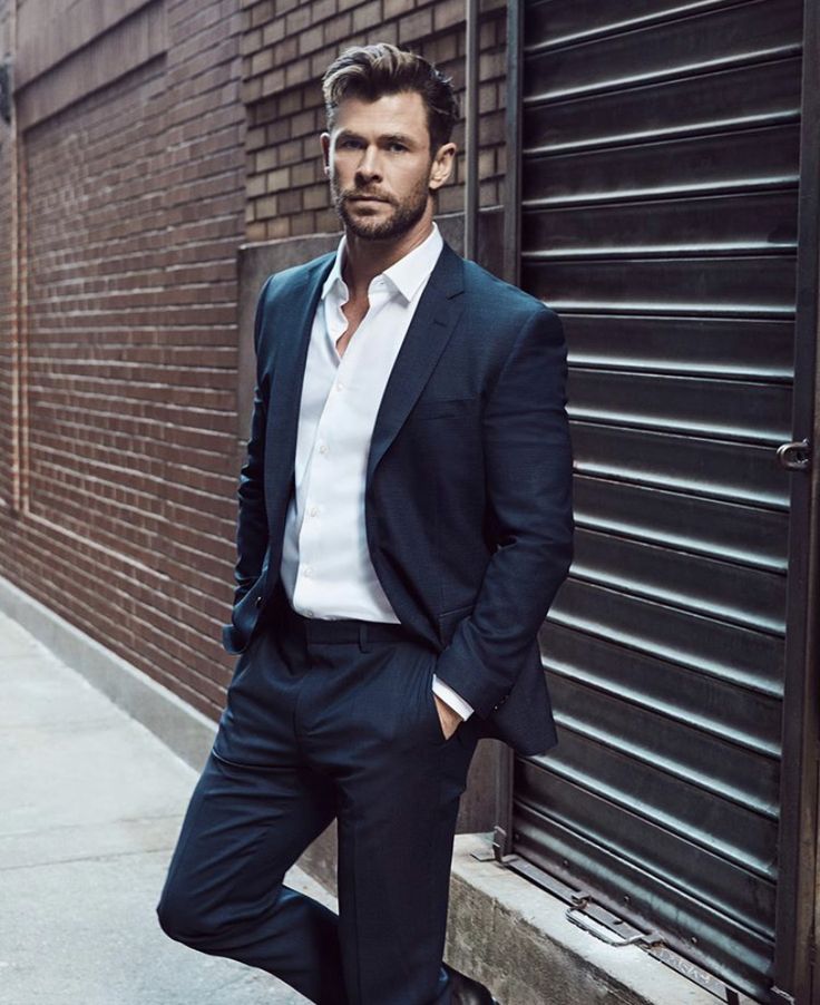 a man in a suit leaning against a brick wall with his hands in his pockets