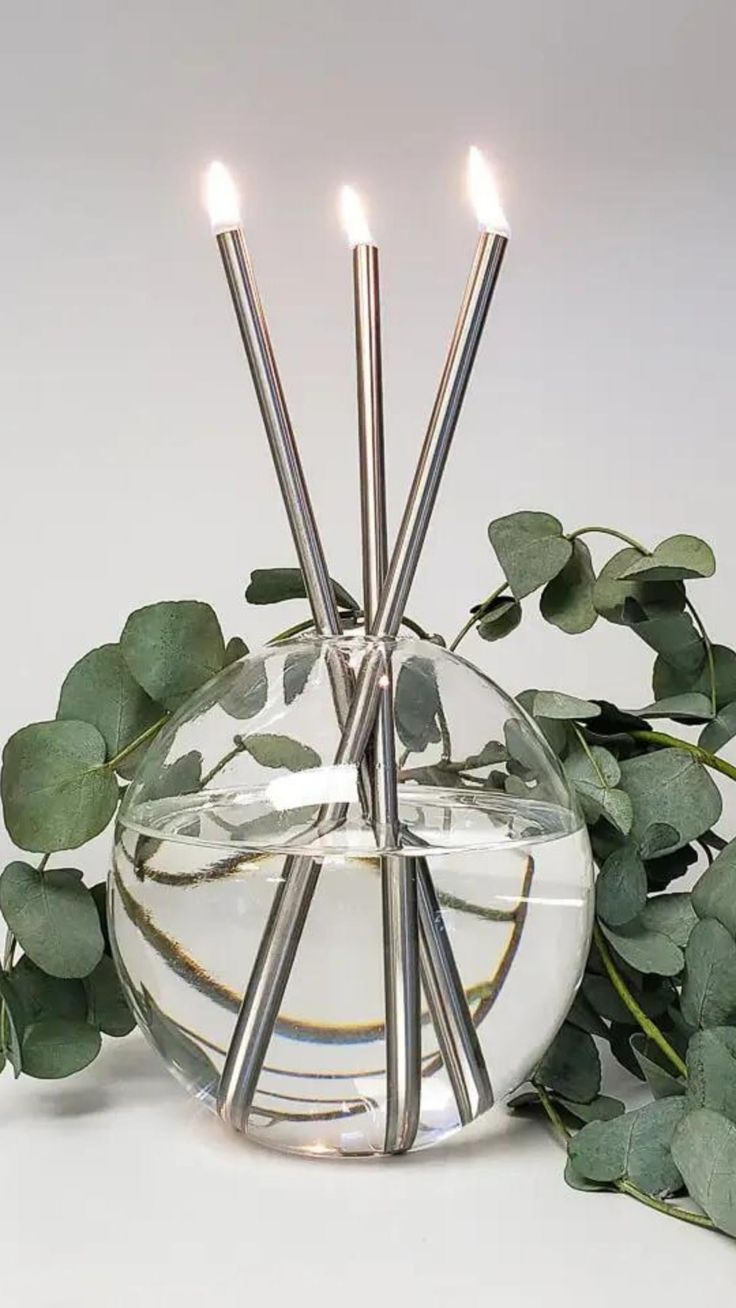 three candles are lit in a glass vase with greenery around it on a white surface