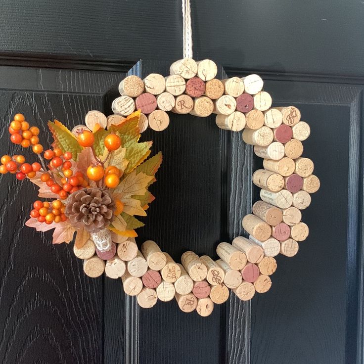 a wreath made out of wine corks hanging on a black door with autumn leaves and berries