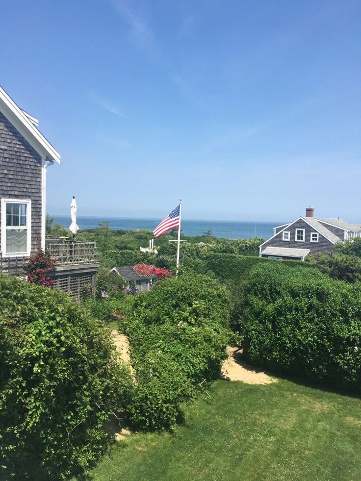 an american flag is flying in front of a house on the ocean side, with trees and shrubs surrounding it