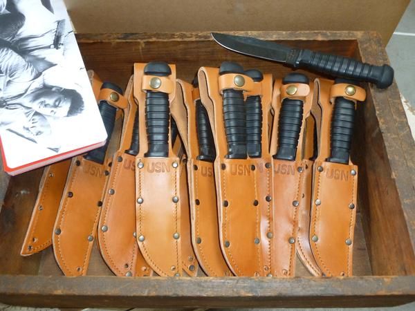 six knives in leather sheaths on a wooden tray with a book and knife sharpener