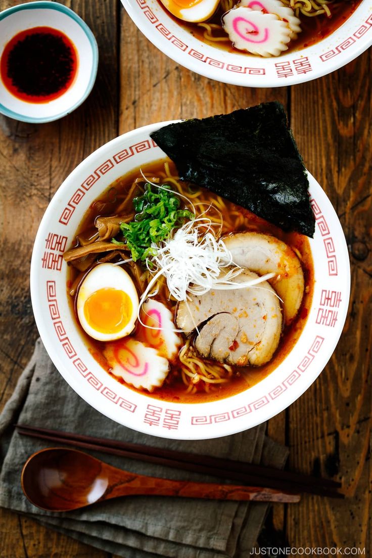 two bowls of ramen with meat, noodles and vegetables on a wooden table next to chopsticks