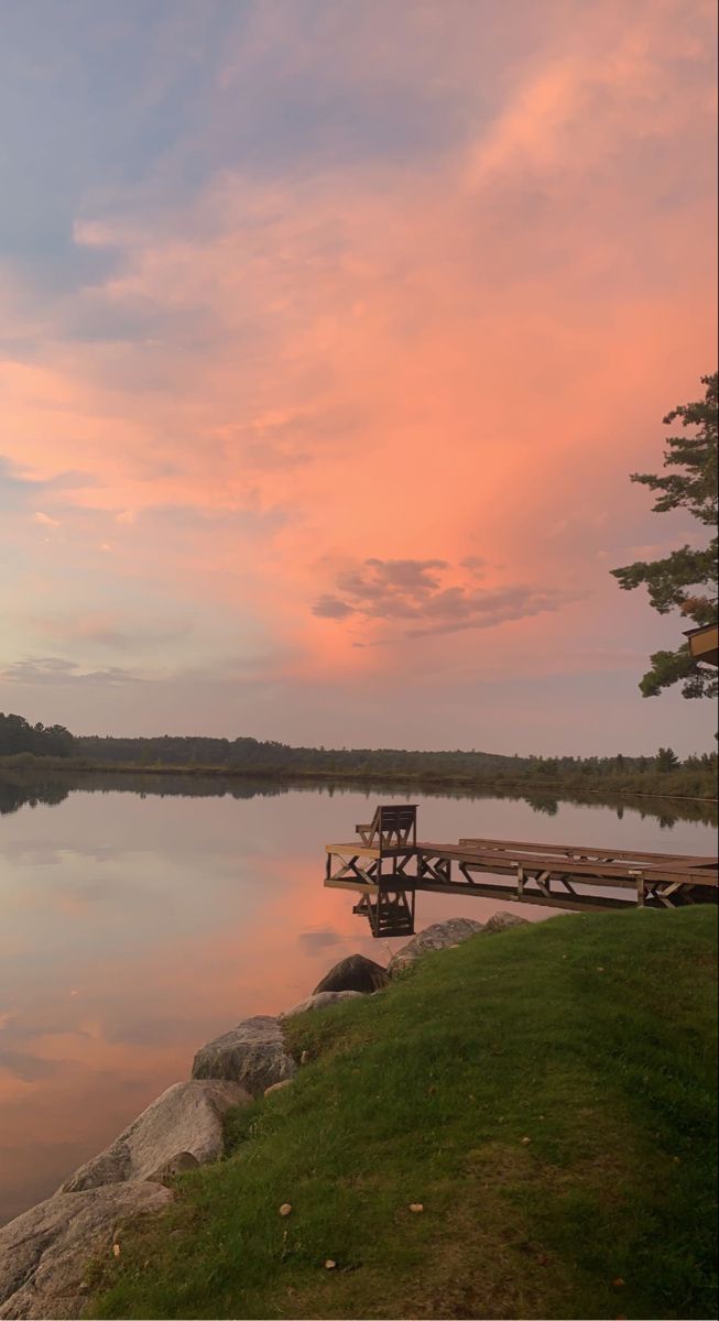 a dock sitting on top of a lush green field next to a lake under a pink sky