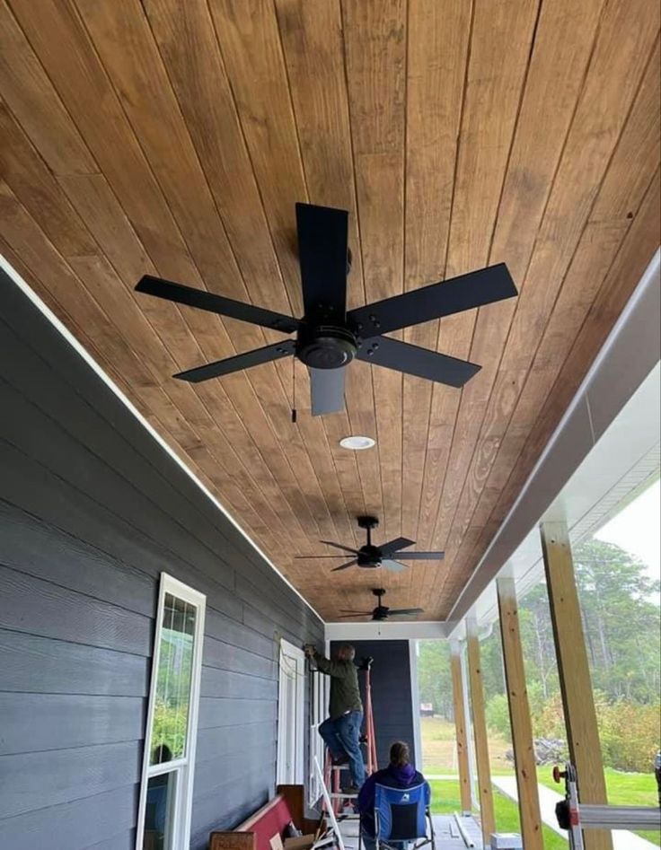 an outdoor covered porch with ceiling fans and chairs on the back deck, along with two people sitting in lawn chairs