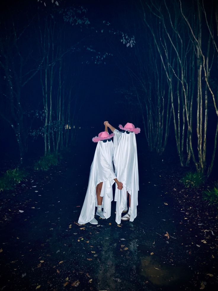 two women in white dresses and pink hats are standing in the dark woods with their arms wrapped around each other