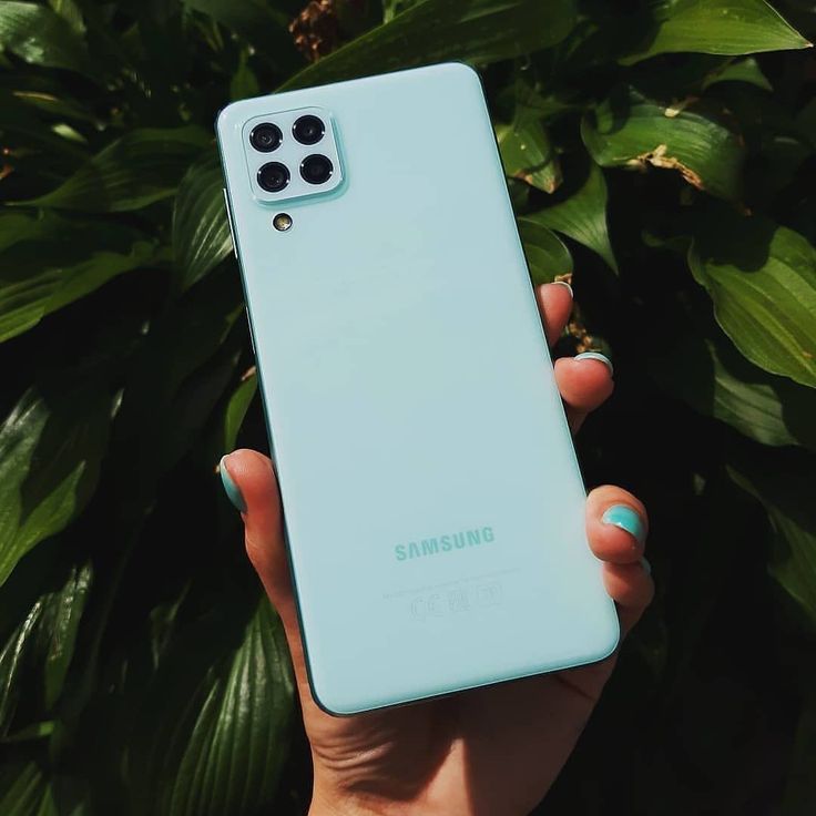 a person holding up a blue samsung phone in front of some plants and trees with green leaves