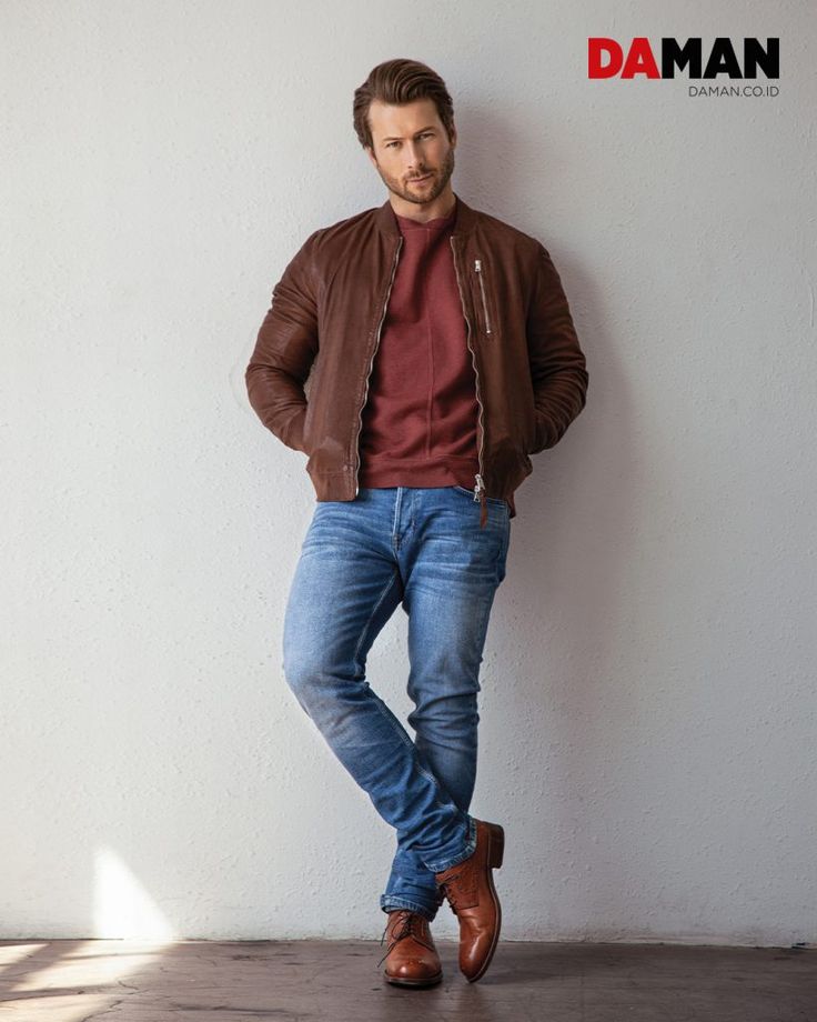 a man leaning against a white wall with his hands in his pockets, wearing jeans and a leather jacket