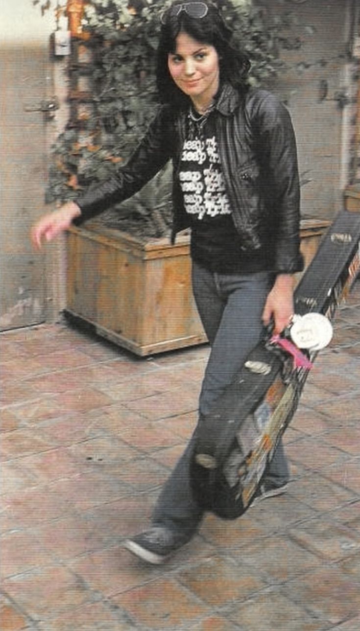 a woman holding a skateboard and wearing a black leather jacket is walking down the street