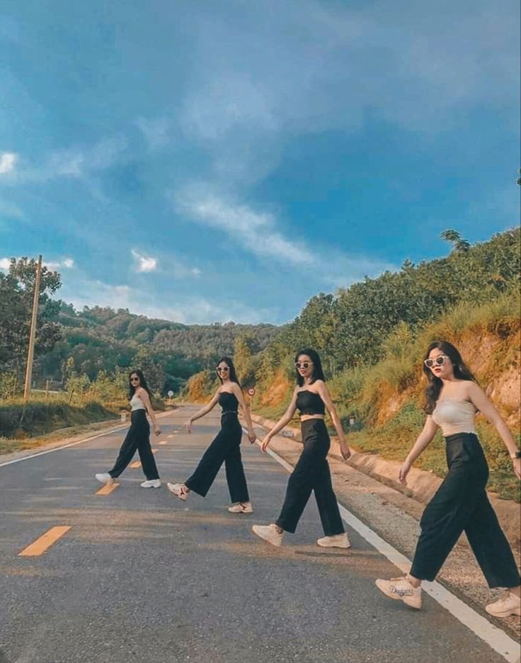 four women in black and white outfits walking across the middle of an empty road with trees on both sides