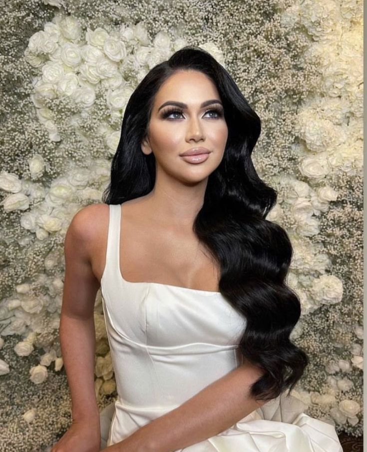 a woman with long black hair sitting in front of a flower covered wall wearing a white dress