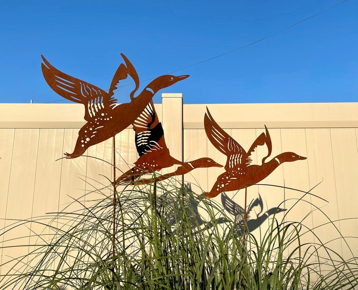 two metal birds sitting on top of tall grass next to a building with a blue sky in the background