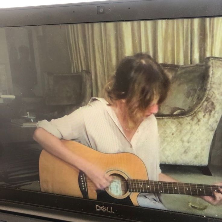 a woman is playing an acoustic guitar in front of a laptop screen with the image of a person sitting on a couch