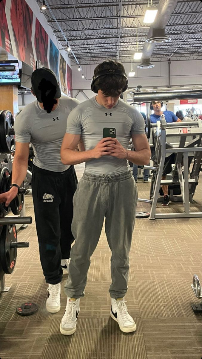 two men standing next to each other in a gym looking at their cell phones while wearing headphones