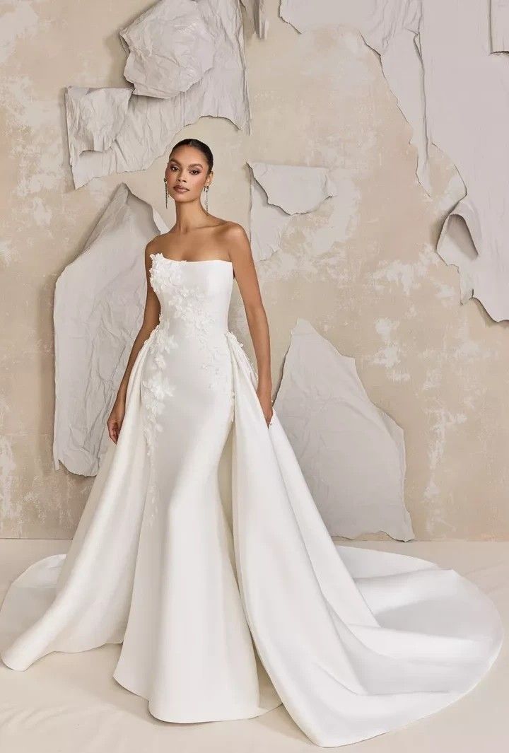 a woman standing in front of a white wall wearing a wedding dress with flowers on it