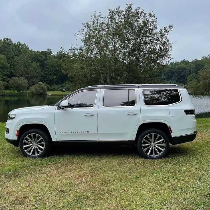 a white suv parked in front of a lake with trees and grass on the side