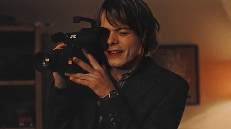 a man holding a camera up to his face in front of a lamp and bookcase