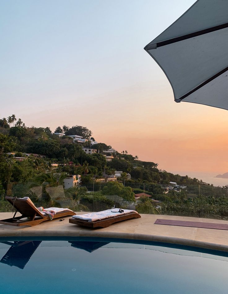 two lounge chairs and an umbrella on the edge of a swimming pool overlooking a hillside