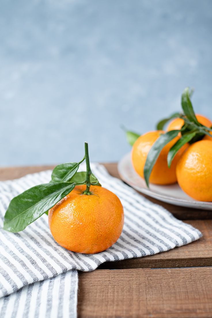 three oranges with leaves sitting on top of a wooden table next to a white plate