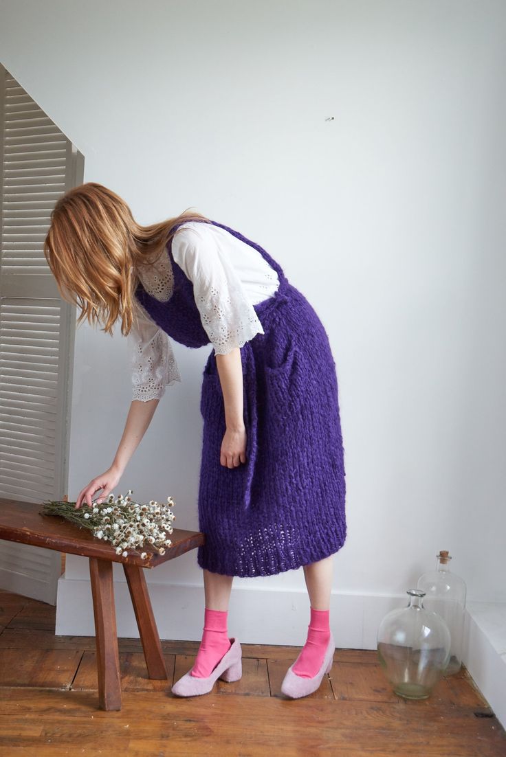 a woman in a purple dress arranging flowers on a small wooden table next to a window